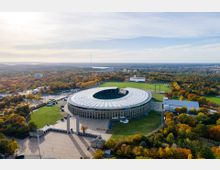 Luftaufnahme eines großen ovalen Stadions mit überdachtem Dach, umgeben von herbstlicher Landschaft mit buntem Laub. Im Vordergrund befinden sich zwei hohe olympische Säulen, und im Hintergrund erstreckt sich eine weitläufige grüne und städtische Umgebung.