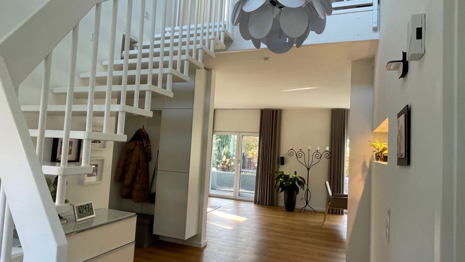 Moderne Wohnraumgestaltung mit einer weißen Treppe, die zu einem offenen Obergeschoss führt. Der Raum ist mit hellen Holzböden, großen Fenstern mit Vorhängen, einem dekorativen Kronleuchter und Pflanzenakzenten eingerichtet.