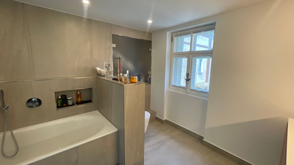 Modernes Badezimmer mit eingelassener Badewanne, grauen Wand- und Bodenfliesen sowie einer Nischenablage in der Wand. Ein großes Fenster sorgt für Tageslicht; daneben befinden sich eine halbhohe Trennwand, ein dunkler Duschbereich und ein WC.