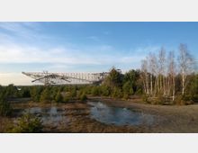 Eine große industrielle Förderbrücke ist in einer offenen Landschaft mit jungen Kiefern und vereinzelten Birkenbäumen zu sehen. Im Vordergrund befinden sich Wasserpfützen auf feuchtem Boden, und der Himmel ist klar mit wenigen Wolken.