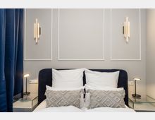 Modernes Schlafzimmer mit dunkel gepolstertem Bett, weißen Kissen und gemusterten Zierkissen vor einer hellgrauen Wand mit Zierleisten und zwei vertikalen Wandleuchten. Links hängen dunkelblaue Vorhänge; neben dem Bett stehen Glas-Nachttische mit schlanken Leselampen.