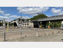 Städtische Straßenszene mit einer erhöhten U-Bahn-Station und einer Fußgängerbrücke, flankiert von einer Statue auf der linken Seite. Im Vordergrund sind Gehwege, gelbe Haltestellenpfosten, parkende E-Scooter und Tauben zu sehen, während Bäume und Gebäude die Umgebung prägen.