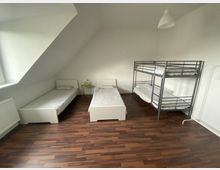 Schlafzimmer mit drei Betten: zwei Einzelbetten mit weißen Rahmen und ein Etagenbett aus Metall. Der Raum hat einen Holzboden, weiße Wände und ein Dachfenster. Ein kleiner Beistelltisch mit einer Pflanze und einer Wasserflasche steht zwischen den Einzelbetten.