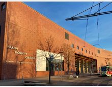 Backstein-Gebäude mit der Beschriftung „Gräfin-Dönhoff-Gebäude, Europa-Universität Viadrina“, ergänzt durch große Fenster und einen überdachten Eingangsbereich. Im Vordergrund verläuft eine Straßenbahnschiene, an der eine Straßenbahn hält, und einzelne Bäume säumen den Gehweg.