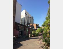 Innenhof in einem umgebauten Industrieareal mit Backstein- und Werkstattgebäuden, großen Fensterfronten und einer bemalten Giebelwand im Hintergrund. Über dem gepflasterten Weg hängen bunte Wimpelketten, daneben stehen Tische und Stühle sowie üppige Grünpflanzen und blühende Sträucher.