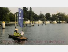 Eine kleine Plattform mit zwei Personen und einem aufblasbaren gelben Boot treibt auf dem Wasser vor einem Hafengebiet. Im Hintergrund sind Boote an Anlegestellen und Bäume zu sehen, während eine blaue Flagge mit der Aufschrift „AHOI“ auffällt.