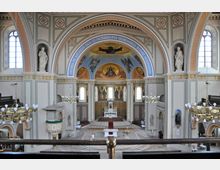 Innenraum einer Kirche mit einem beeindruckenden Altarbereich, geschmückt mit Ikonen und goldenen Elementen. Hohe Rundbögen mit farbigen Verzierungen, Wandgemälde über dem Altar und Kronleuchter unterstreichen die dekorative Architektur.