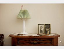 Holzkommode mit einer Tischlampe mit grünem, gefaltetem Lampenschirm, zwei gerahmten Schwarz-Weiß-Fotografien und einem Löffel auf einer weißen Tischdecke. Im Hintergrund eine hellbeige Wand mit kleinen Farbflecken.