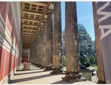 Säulenhalle mit rötlich gestalteten Wandpaneelen und Kassettendecke, die Blick auf den Berliner Dom im Hintergrund freigibt. Rechts steht eine Bronzeskulptur, und der Außenbereich zeigt grüne Bäume und Passanten.