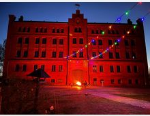 Rotes Backsteingebäude mit mehreren Fenstern, beleuchtet von farbigen Lichterketten und einem Feuer im Vordergrund; nächtliche Szenerie mit gepflastertem Vorplatz und dunklem Himmel.