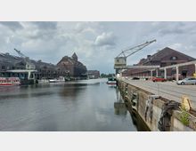 Industriehafen mit Backsteingebäuden auf beiden Seiten des Kanals, ausgestattet mit alten Hafenkränen. Auf dem Wasser liegen mehrere Boote vor Anker, während Autos auf einer gepflasterten Straße entlang des Ufers parken.
