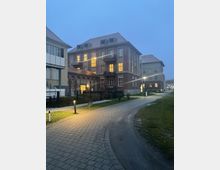 Mehrstöckiges Backsteingebäude mit beleuchteten Fenstern am Abend, umgeben von einem gepflasterten Weg, Rasenflächen und seitlich stehenden Straßenlaternen; im Hintergrund weitere ähnliche Gebäude.