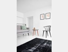 Minimalistisch eingerichtetes Wohnzimmer mit einem grauen Sofa, einem kleinen runden Tisch mit Holzbeinen, einer schwarzen Designerstuhl und zwei gerahmten Bildern an der weißen Wand. Der Boden ist weiß, und ein dunkler Teppich liegt im Vordergrund.