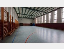 Leere Sporthalle mit großem Hallenboden und farbigen Spielfeldmarkierungen, an den Seiten mit Sprossenwänden und Bänken. Hohe Fensterreihen lassen viel Tageslicht herein, darüber eine Decke mit sichtbaren Trägern und Leuchten.