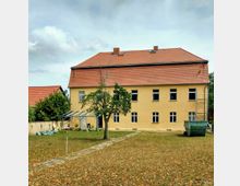 Gelbes zweistöckiges Gebäude mit rotem Ziegeldach, mehreren symmetrischen Fenstern und einem kleinen Glasdach-Anbau an der linken Seite. Im Vordergrund ein Rasenbereich mit einem gepflasterten Weg, einem einzelnen Baum und einem grünen Baucontainer.