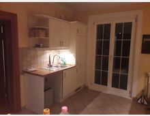 Eine kleine Küche mit cremefarbenen Schränken, einer Spüle mit Metallarmatur und einer Holzarbeitsplatte. Rechts befindet sich eine verglaste Doppeltür mit Sprossenrahmen, daneben beige Fliesen und gelb gestrichene Wände.