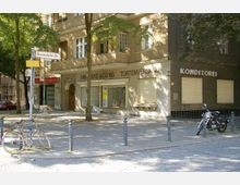 Ecke eines Wohn- und Geschäftshauses mit der Beschriftung "P. Mieske & Co KG Tortenversand Konditorei". Der Gehweg ist mit Kopfsteinpflaster bedeckt, daneben stehen ein Fahrrad und ein Motorrad, und es gibt vereinzelte Bäume, die Schatten werfen.