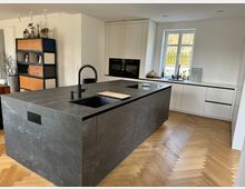 Moderne Küche mit einer dunklen Kücheninsel aus Steinoptik, integriertem Spülbecken und Wasserhahn, umgeben von weißen Schränken mit minimalistischen Griffen. Der Raum hat einen hellen Fischgräten-Holzboden, ein großes Fenster mit Blick ins Grüne und ein Regal mit Holz- und Metallakzenten an der Seitenwand.