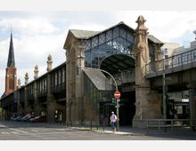 Straßenansicht eines historischen Bahnhofs in Berlin mit einer erhöhten Gleisanlage und einem überdachten Eingangsbereich aus Glas und Stein. Im Hintergrund ist der Turm einer Backsteinkirche mit einer Uhr zu sehen.