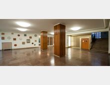 Ein heller Eingangsbereich mit poliertem Steinboden, zwei hölzernen Säulen und einer Wand mit zahlreichen Uhren unterschiedlicher Designs. Rechts befindet sich eine Treppe und eine Tür, links ein Heizkörper unter der Uhrensammlung.