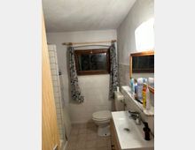 Kleines Badezimmer mit hellen Fliesen an Wänden und Boden. Es gibt ein kleines Fenster mit bunten Vorhängen, eine Toilette, ein Waschbecken mit Spiegel darüber und verschiedene Hygieneartikel auf einer Ablage.