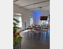 Ein kleiner Raum mit weißen Wänden und gewölbter Decke, ausgestattet mit mehreren Holzstühlen in Reihen. Große Fenster lassen Tageslicht herein, und Pflanzen sowie ein Bildschirm sind im Hintergrund sichtbar.