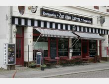 Eine Straßenansicht eines Restaurants mit dem Namen „Zur alten Laterne“. Das Gebäude hat weiße Wände, rot gerahmte Fenster und Türen, gestreifte Markisen sowie eine Außenterrasse mit roten Tischen und grünen Stühlen; daneben befindet sich ein Plakatkasten mit der Aufschrift "THEATER".