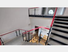 Ein modernes Treppenhaus mit Betonstufen, Glasgeländern und roten Handläufen. Im Untergeschoss befinden sich Holzmöbel und eine Garderobe vor einer weißen Wand.