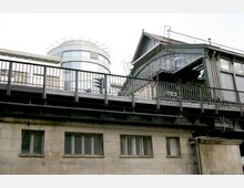 Außenansicht einer erhöhten U-Bahn-Station mit Metallstruktur und Geländern, darunter ein steinernes Gebäude mit rechteckigen Fenstern. Im Hintergrund befindet sich ein moderner, verglaster Turm.