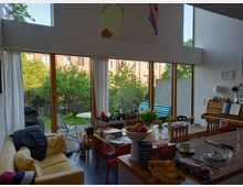 Moderne Wohnküche mit großen, bodentiefen Fenstern, die einen Blick auf einen Garten mit Bäumen und Gartenmöbeln freigeben. Im Innenraum befinden sich ein Esstisch, Sofa, Klavier und Küchentheke, dekoriert mit Obst und Blumen.