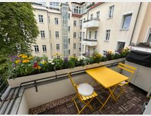 Blick von einem Balkon in einen Innenhof mit hellen Mehrfamilienhäusern, vielen Fenstern, kleinen Balkonen und einem gläsernen Aufzugschacht. Im Vordergrund stehen ein gelber Klapptisch mit zwei Stühlen auf Holzfliesen sowie Blumenkästen mit bunten Blumen entlang des Geländers.
