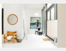 Helles, modernes Wohnzimmer mit weißem Boden und großen bodentiefen Fenstern. Im Vordergrund stehen ein Ledersessel mit Fellauflage, ein runder Holzspiegel an der Wand und eine Stehlampe, im Hintergrund ist ein Gemälde vor einer weißen Wand sichtbar.