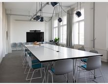 Heller, moderner Besprechungsraum mit langen weißen Konferenztischen und grauen Stühlen, darüber hängende schwarze Pendelleuchten und sichtbare Deckeninstallationen. Große Fenster mit Vorhängen lassen viel Tageslicht herein; an der Stirnseite steht ein großer Bildschirm.