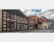 Straße mit historischen Fachwerkhäusern, gekennzeichnet durch weiße Wände mit roten Balken, Holzdetails und teils farbigen Fensterläden; daneben stehen Putzgebäude in Pastellfarben. Die Häuser grenzen direkt an den Gehweg, unter einem bewölkten Himmel.