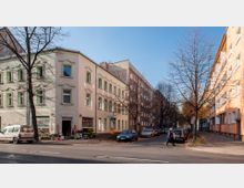 Straßenecke in einem Wohngebiet mit mehrstöckigen Gebäuden aus hellem Putz, Bäumen ohne Laub und parkenden Autos entlang der Straße. Im Vordergrund ein Laden mit Blumen, ein Gehweg, und ein Fußgänger, der die Straße überquert.