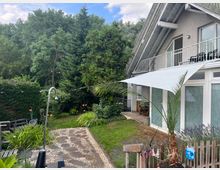 Ein modernes Einfamilienhaus mit hellgrauer Fassade und großem Balkon, umgeben von einem gepflegten Garten mit Bäumen, Sträuchern und Blumen. Im Vordergrund befindet sich ein gepflasterter Weg, ein kleiner Zaun und eine Außendusche.