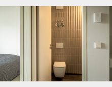 Ein modernes Badezimmer mit einer Wand aus strukturierten, beige-braunen Fliesen, einer wandmontierten Toilette und einer teilweise geöffneten weißen Tür. Im Vordergrund ist ein angrenzender Raum mit einem Bett und einer schwarz-weiß gemusterten Bettdecke sichtbar.