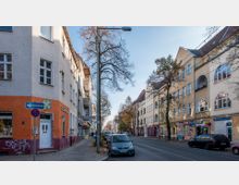 Städtische Straße mit mehrstöckigen Wohnhäusern auf beiden Seiten, teils mit farblich unterschiedlichen Fassaden und großen Fensterbögen. Im Vordergrund geparkte Autos, Gehwege, Bäume mit Herbstlaub sowie Straßenschilder, darunter ein Schild für eine Einbahnstraße.