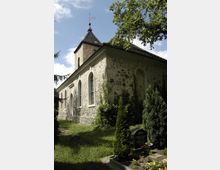 Steinbaukirche mit einfachem Glockenturm und schrägem Dach, umgeben von Grünflächen, Bäumen und einem kleinen Friedhof mit Grabstein und Blumen im Vordergrund.