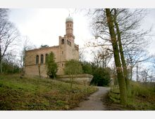 Backsteingebäude mit Kuppelturm, umgeben von Bäumen, auf einem leicht erhöhten Gelände. Ein mit Geländern gesäumter Weg führt durch das Grün auf das Gebäude zu.