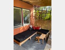 Eine überdachte Terrasse mit einer orangen Wand und einem Bambusdach. Es gibt zwei Holzbänke mit schwarzen Polstern, roten und weißen Kissen, einen kleinen Holztisch und eine Hängeleuchte mit lilafarbenen Lampenschirmen. Im Hintergrund sind Pflanzen mit einem Gitter aus Holz zu sehen.