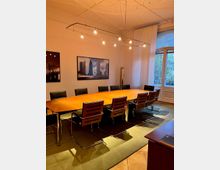 Konferenzraum mit einem langen Holztisch, umgeben von schwarzen Stühlen mit Chromgestell. An der Wand hängen Bilder von Stadtansichten, und durch bodentiefe Fenster fällt Tageslicht herein. Eine moderne Deckenbeleuchtung und ein Teppich runden den Raum ab.