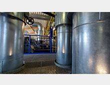 Industrieanlage mit großen metallischen Rohren und Tanks in einer Halle mit Wellblechdach. Im Hintergrund sind blaue Geländer, gelbe Leitungen und technische Geräte sichtbar, die auf eine industrielle Umgebung hinweisen.