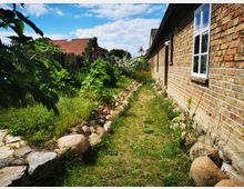 Schmaler Gartenpfad neben einer Backsteinmauer mit weißen Fenstern, gesäumt von Gras, Blumen und Steinen; im Hintergrund ein Haus mit rotem Ziegeldach und ein Holzzaun unter einem blauen Himmel.