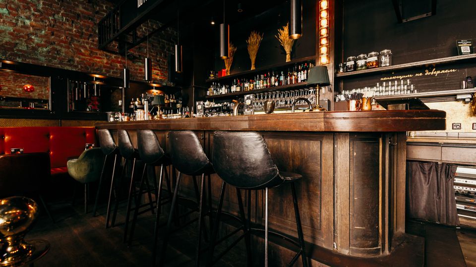 Innenansicht einer Bar im Industrial-Stil mit langer dunkler Holztheke, mehreren hohen Barhockern und Regalen voller Flaschen und Gläser. Sichtmauerwerk aus Ziegeln, schwarze Wände und hängende Zylinderleuchten prägen den Raum.