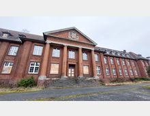 Backsteinbau mit symmetrischer Fassade, großen Fenstern und mehreren Säulen im Eingangsbereich. Ein Treppenaufgang führt zum Haupteingang, teilweise mit Holzplatten verbarrikadierte Fenster und verwilderte Pflanzen umgeben das Gebäude.