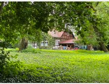 Fachwerkhaus mit rotem Ziegeldach, teilweise von Efeu bewachsen, umgeben von Bäumen und einer grünen Wiese; vor dem Haus stehen ein blauer Traktor und einige Gegenstände.