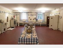 Große historische Küche mit weißen Fliesen an den Wänden, einem roten Fliesenboden und einem zentralen Tisch mit blau-weiß-karierter Tischdecke, auf dem Geschirr und eine Teekanne arrangiert sind. Im Hintergrund befinden sich hohe Fenster mit Vorhängen, Schränke und Wanddekorationen mit Keramikkacheln.