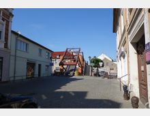 Straßenszene in einer Altstadt mit Kopfsteinpflaster, flankiert von mehrstöckigen Gebäuden. Im Hintergrund eine Fußgängerbrücke mit Metallkonstruktion und Treppen, umgeben von historischen und modernisierten Häusern.