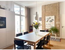 Heller Ess- und Küchenbereich mit großem rechteckigem Esstisch und mehreren schwarzen Stühlen, neben hohen Fensterfronten mit Vorhängen. An der Wand befindet sich eine freigelegte Ziegelwand mit Poster, Pflanzen und Hängeleuchte; links sind weiße Einbauschränke mit Backofen.